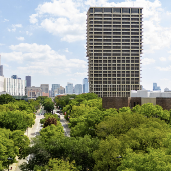 Tall building (grey and tan), green trees in forefront, clouds in background